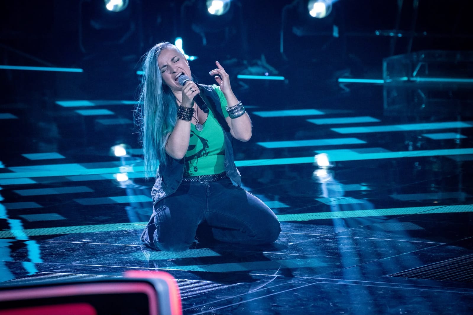 Biggi Käfer kniet singend auf der Bühne von The Voice of Germany, türkise Haare, petrolblaues Bühnenlicht